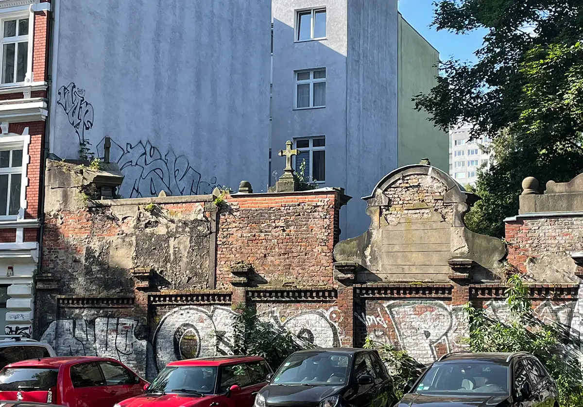 Rückseitige Friedhofsmauer mit brüchigen Steinen und Steinkreuz, davor parkende Autos