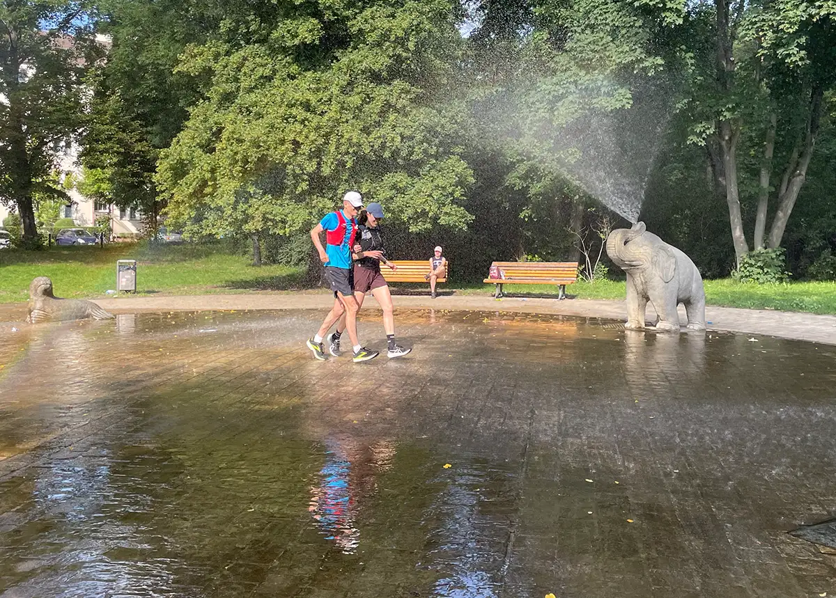 Läuferin und Läufer gehen unter einem sprühenden Wasserstrahl hindurch, der aus dem Rüssel eines Stein-Elefanten kommt