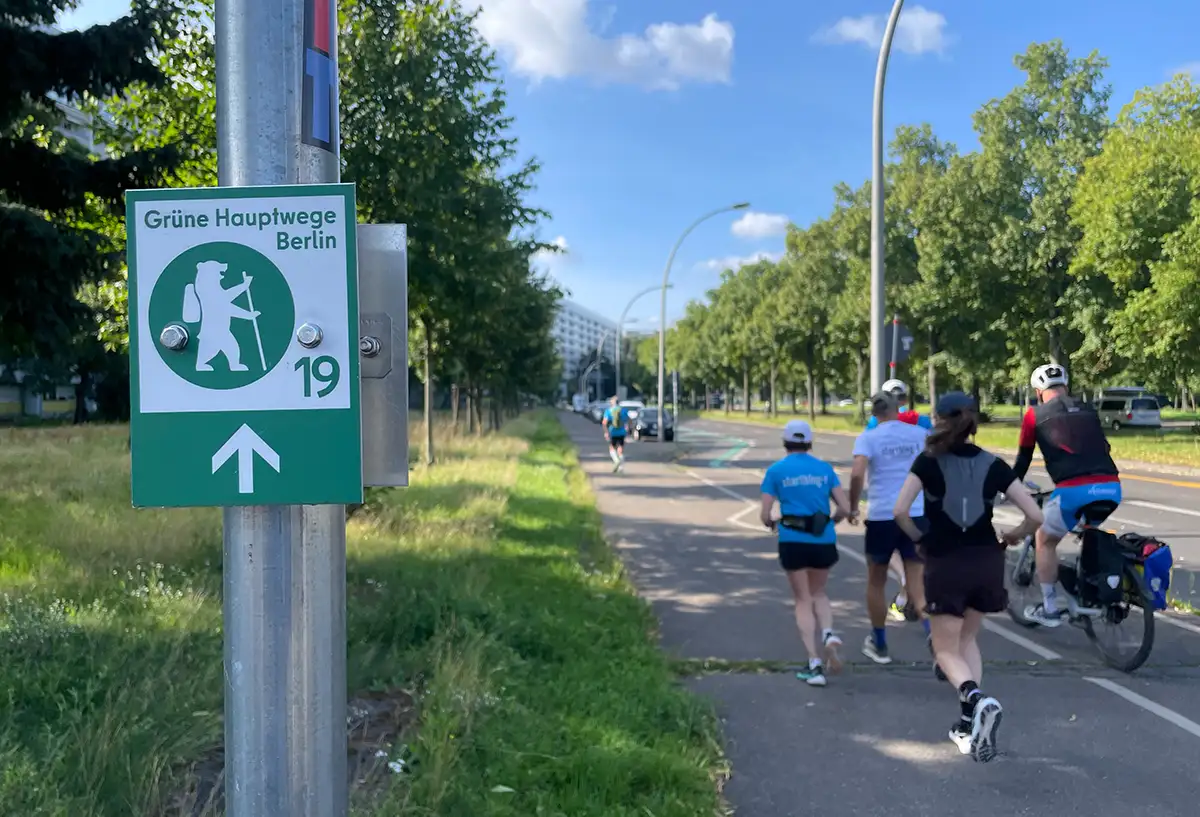 Im Vordergrund ein Schild an einem Metallmast: Grüne Hauptwege Berlin 19, daneben die Laufgruppe auf dem Fußweg einer Straße