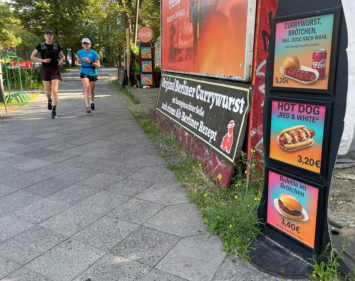 Läuferinnen auf einem Gehweg, im Vordergrund Werbeschilder einer Currywurstbude