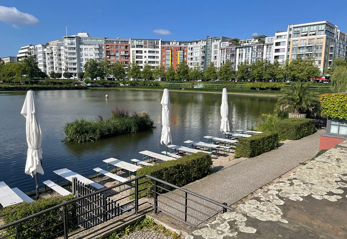 Engelbecken mit weißen Holzbänken und -tischen im vordergrund und Wohnbebauung im Hintergrund