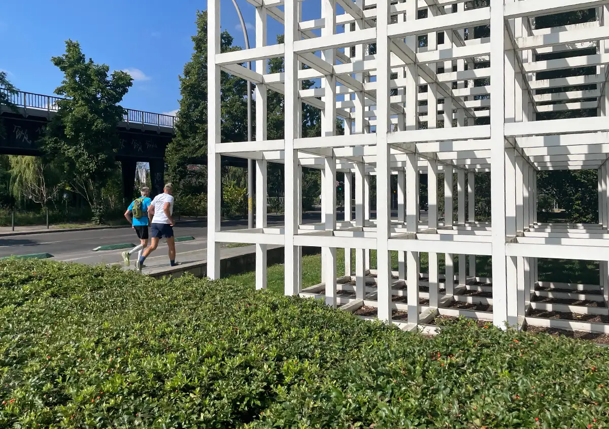 2 Läufer passieren eine moderne Skulptur mit weißen Streben, die viele kleine Kuben in einem großen Kubus bilden