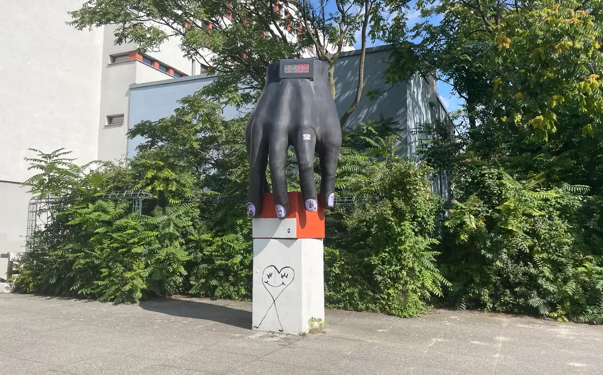 Skulptur „Hand mit Uhr“: eine große bronzene Hand fasst einen orangefarbenen Würfel, in die Hand ist am Handgelenk eine Digitaluhr eingelassen, die gerade 11:00 anzeigt