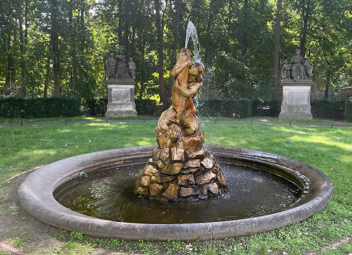 Goldener Tritonbrunnen am Rande des Tiergartens: Der Meeresgott kniet und hält einen großen Fisch aus dem das Wasser des Brunnens sprießt