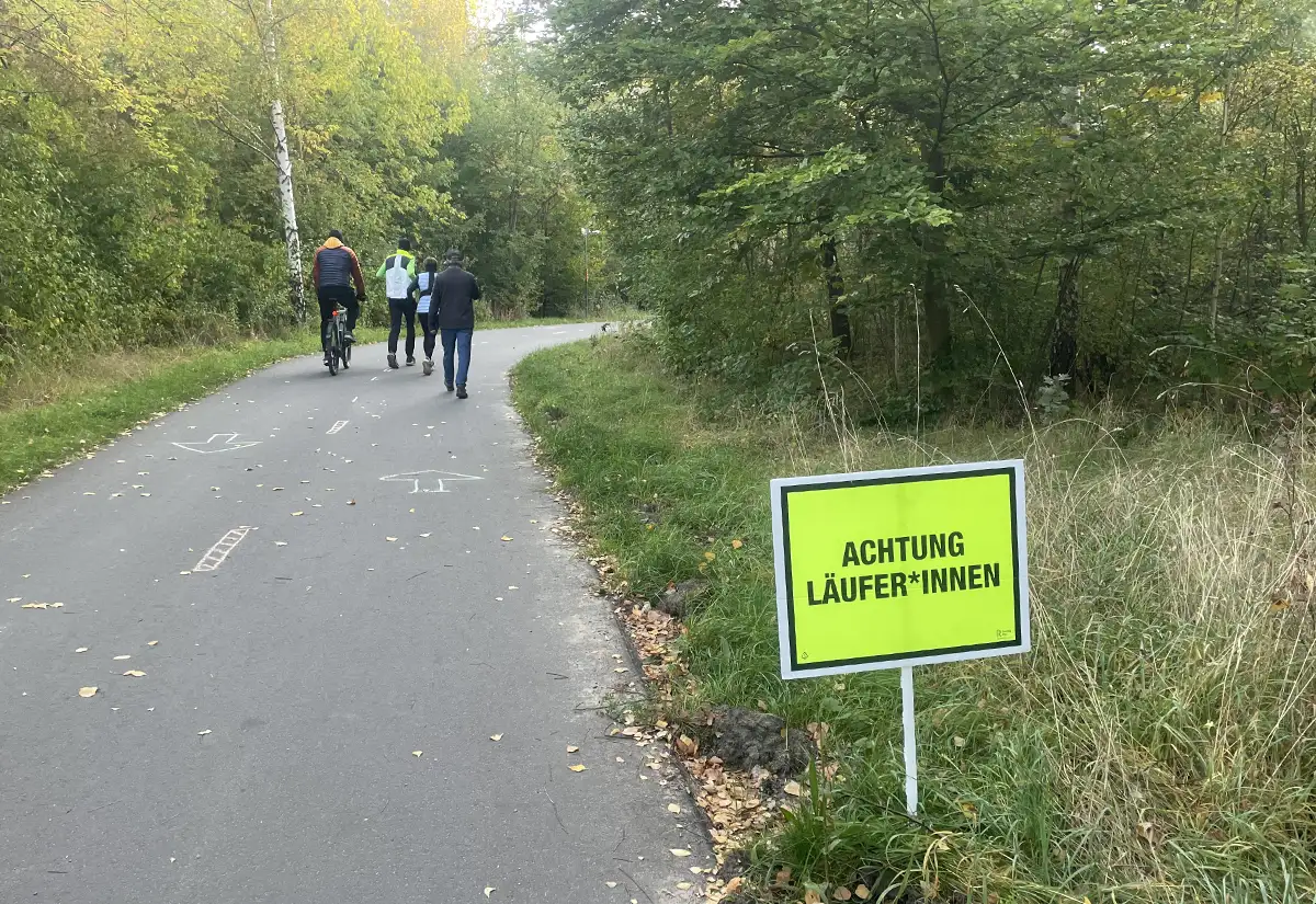 Laufgruppe auf dem Berliner Mauerweg, im Vordergrund ein Schild „Achtung Läufer*innen“