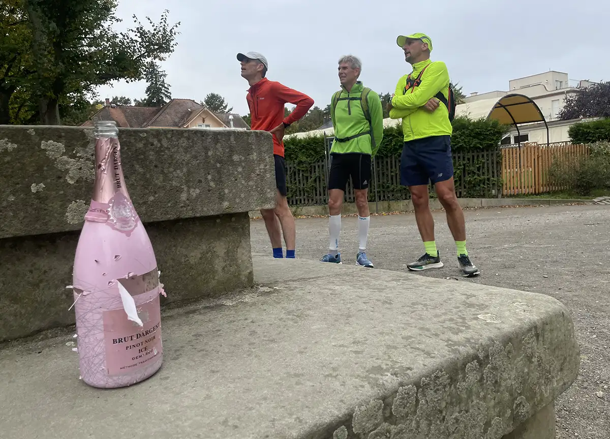 3 Läufer lesen die Beschriftung am Denkmal, im Vordergrund steht eine rosa Sektflasche auf den Stufen
