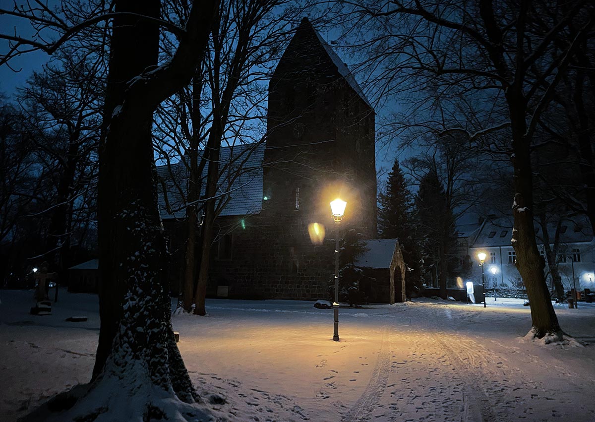 Alte Dorfkirche mit verschneiten Wegen in der Morgendämmerung, davor eine hell strahlende Laterne