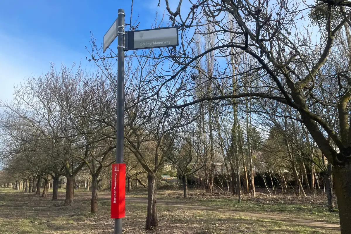 Schild Berliner Mauerweg, dahinter noch kahle Kirschbäume