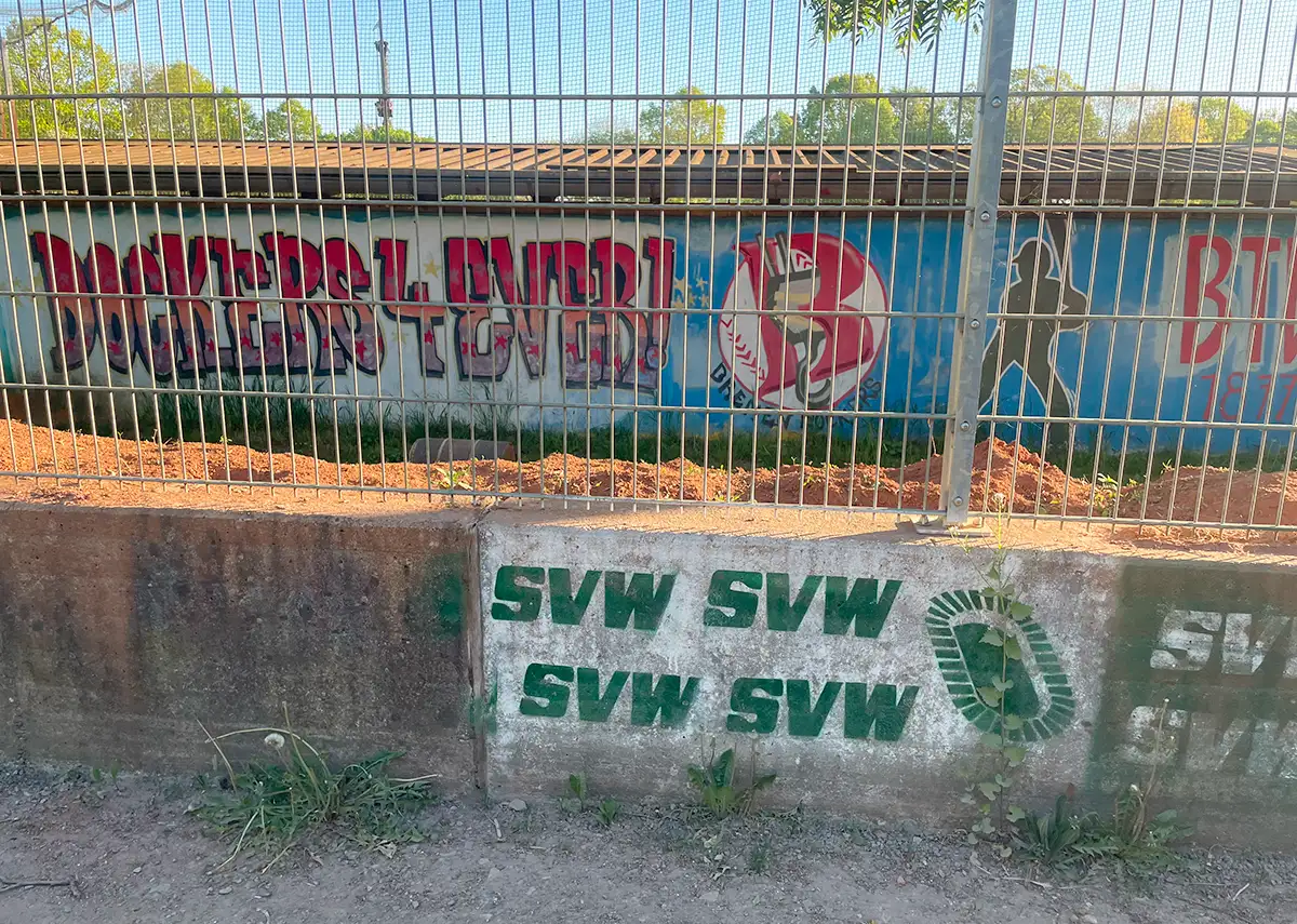 Mauer mit Metallzaun und Aufschrift SVW, dahinter Gebäude mit Graffiti Dockers4Ever und Baseball-Figur