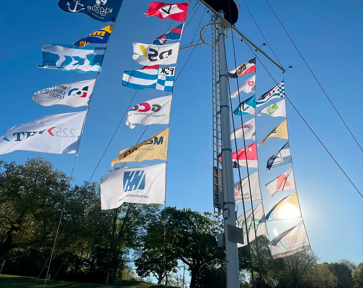 Viele Firmen-Fahnen an einem großen Flaggenmast im Gegenlicht