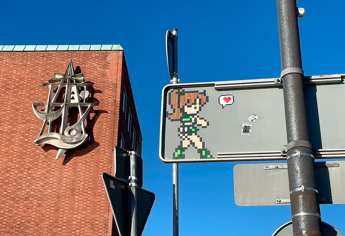 Skulptur an einem Gebäude, im Vordergrund die Rückseite eines Schildes auf dem eine Pixelfigur mit Herz aufgemalt ist