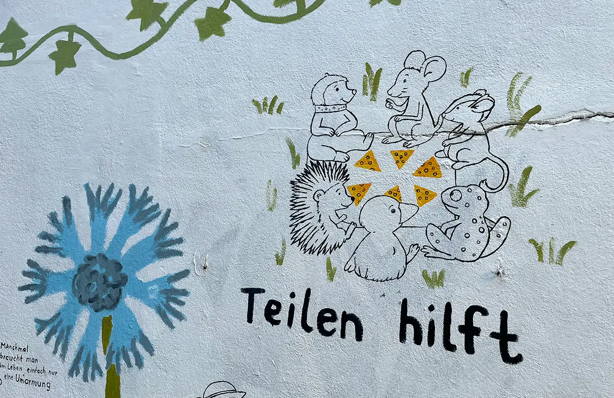 Bemalte Hauswand mit Zeichnung kleiner Tiere, die Essen teilen und Beschriftung „Teilen hilft“