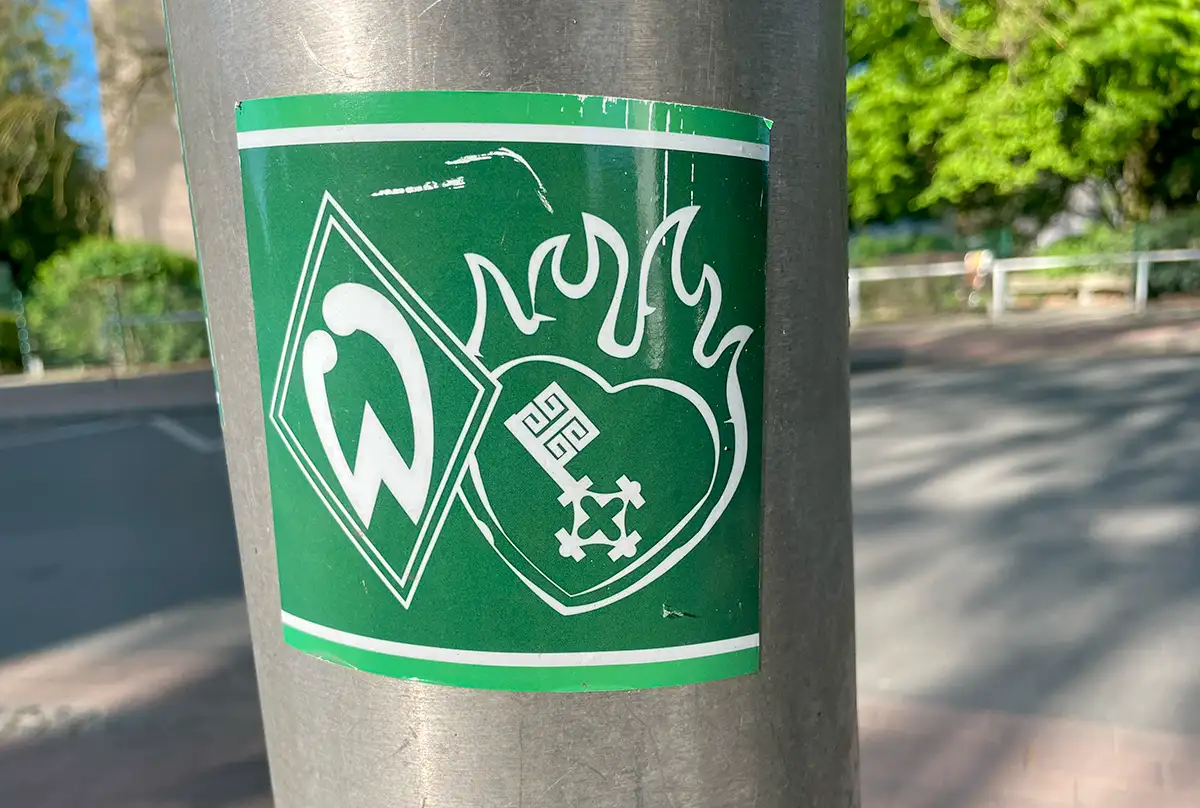 Grüner Aufkleber an einem Laternenmast mit Werder-Logo und brennendem Herz mit Bremer Schlüssel