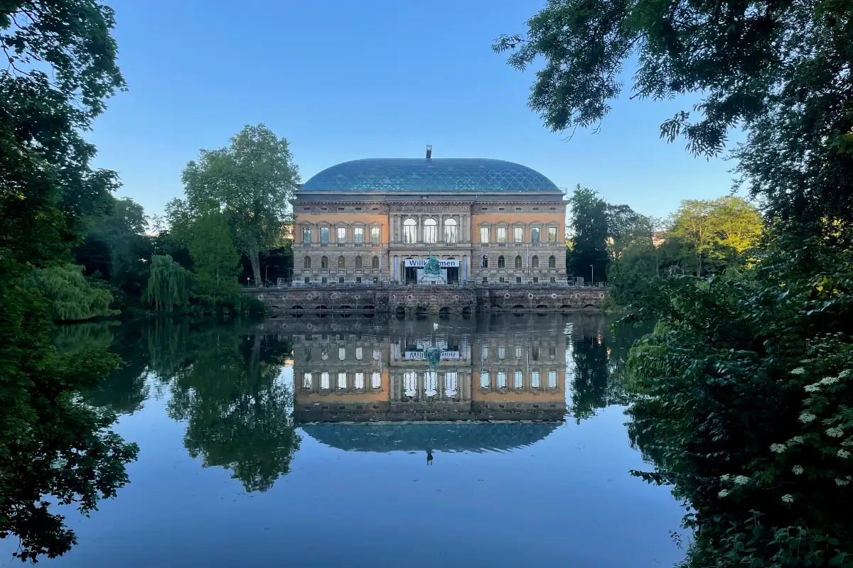 Großes klassizistisches Gebäude an einem See, in dem sich Gebäude und Bäume spiegeln