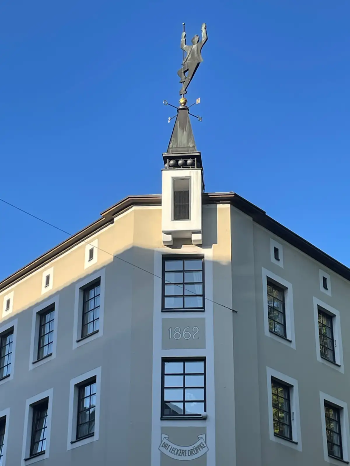 Hausecke mit einem an der Stange hängenden Mann als Wetterfahne, am Haus steht „Dat leckere Dröppke“