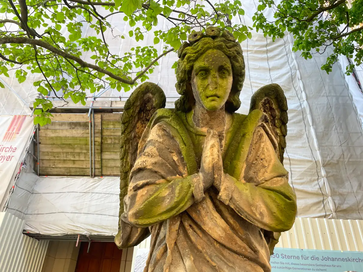 Verwitterte Stein-Skulptur eines Engels vor einer eingerüsteten Kirche