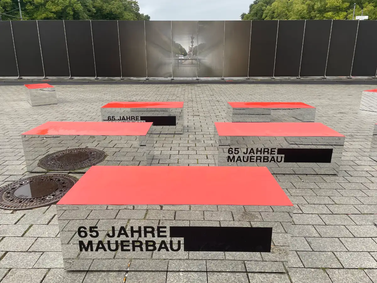 Spiegelnde Bänke mit Aufschrift 65 Jahre Mauerbau, dahinter die Kunst-Mauer-Installation