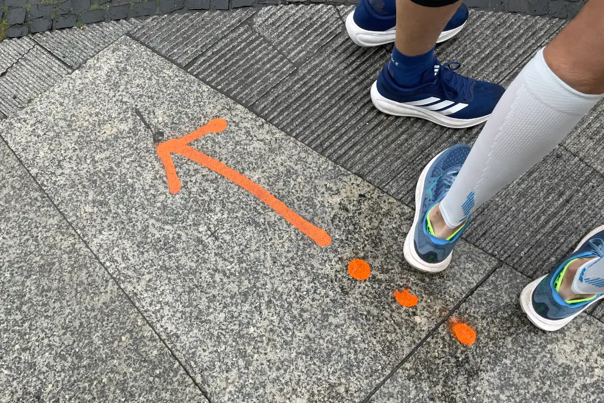 Läuferbeine, auf dem Asphalt ein oranger Pfeil mit drei Punkten